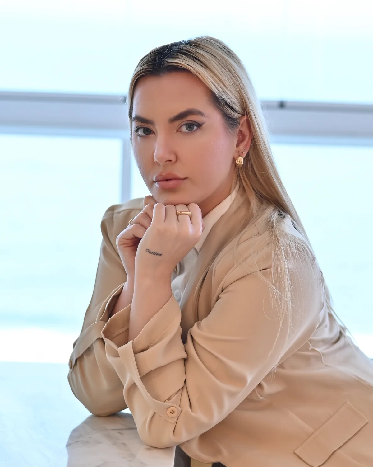 Graziele Dalpra de bege, apoiada na mesa com as mãos no queixo, diante de uma janela clara